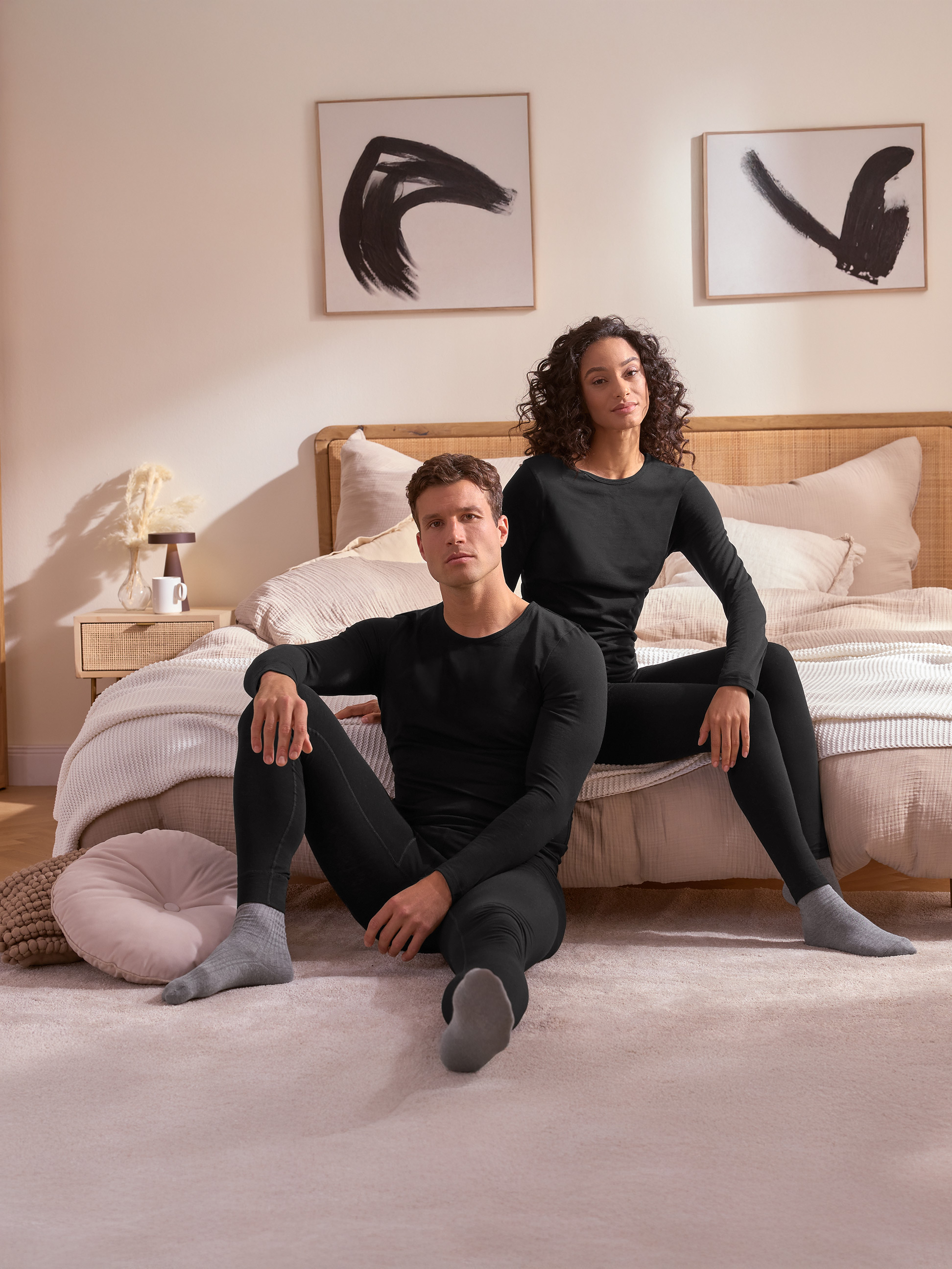 Un couple en sous-vêtements thermiques noirs assis sur le sol devant un lit.