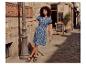 Femme en robe d'été bleue et sandales noires, posant dans une rue pavée.