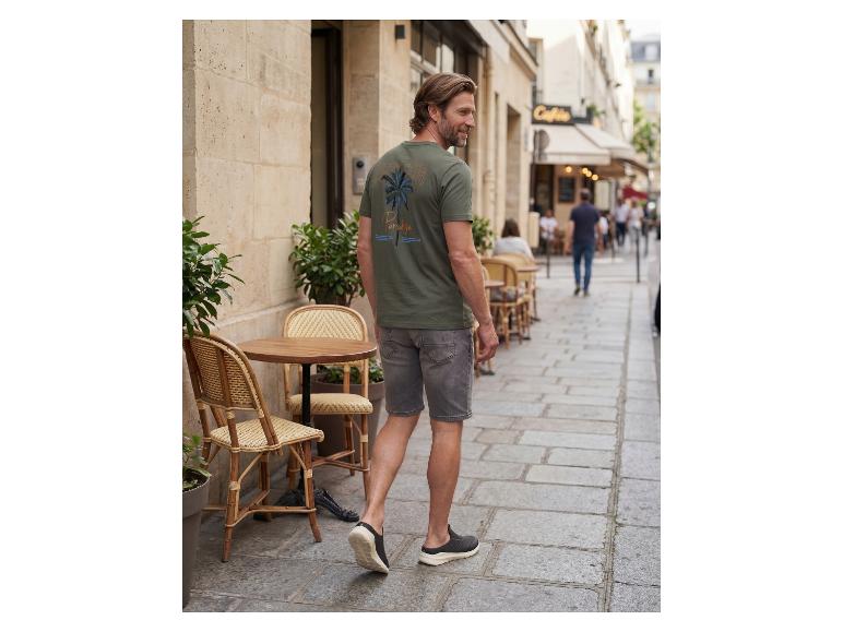 Man in groen T-shirt met palmboomprint en grijze korte broek loopt over een straat.