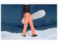 Een vrouw in een oranje skibroek met een snowboard en een bergachtig landschap op de achtergrond.
