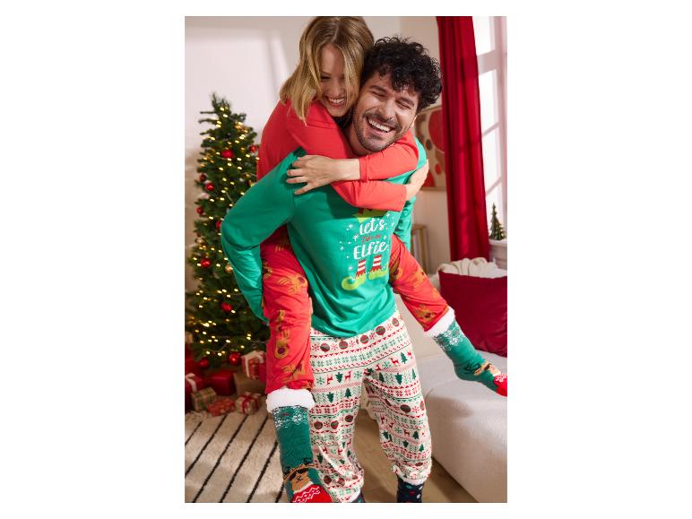 Gelukkig stel in kerstpyjama's, man draagt vrouw op zijn rug naast een kerstboom.