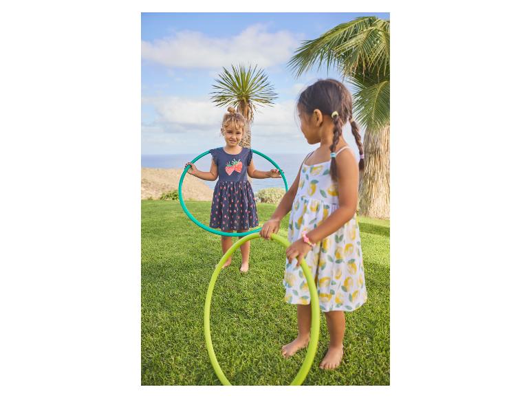 Twee meisjes spelen met hoepels op het gras, met palmbomen en de zee op de achtergrond.