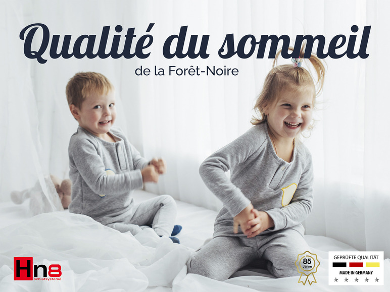 Deux enfants en pyjama gris sur un lit blanc, publicité pour les produits de sommeil Hne Schlafsysteme.