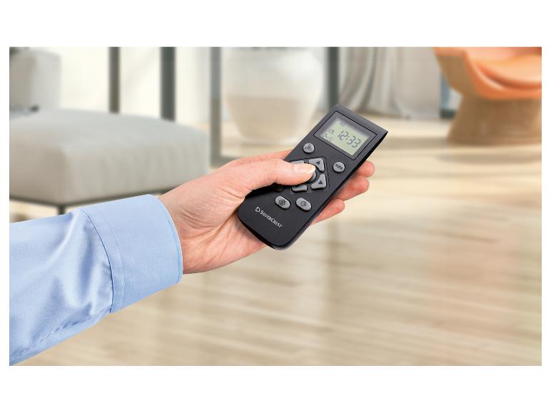 Een hand houdt een zwarte afstandsbediening vast met een digitaal display dat 12:33 toont, in een lichte kamer.