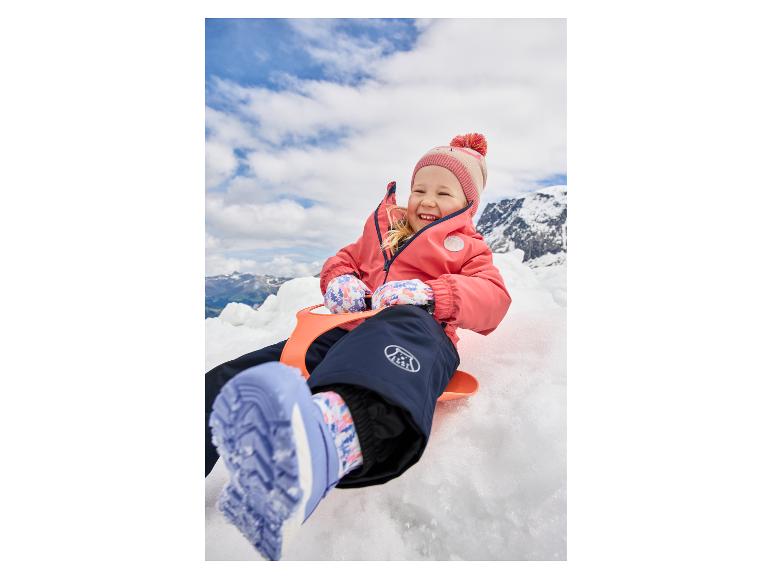 Fillette souriante en veste rose et pantalon de neige bleu, assise sur une luge orange dans la neige.