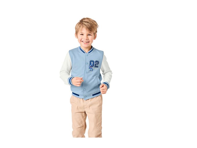 Lachende jongen in een lichtblauwe en witte varsity-jas met een stripfiguur en een beige broek.