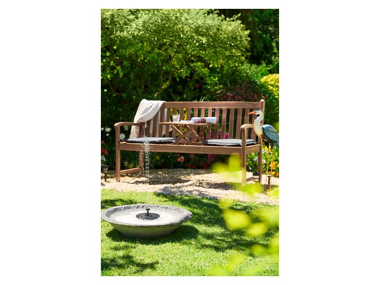 Banc de jardin en bois avec petite table, coussins et fontaine pour oiseaux dans un jardin ensoleillé.