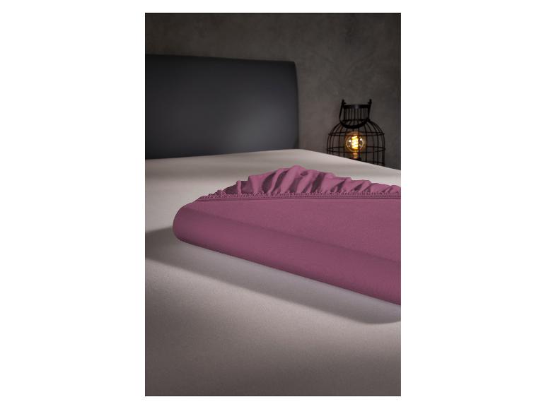 Opgevouwen paars hoeslaken op een bed met grijs hoofdbord en decoratieve lamp.