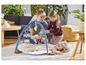 Un bébé est allongé sur un tapis de jeu avec un arc de jeu avec des jouets, deux enfants regardent.