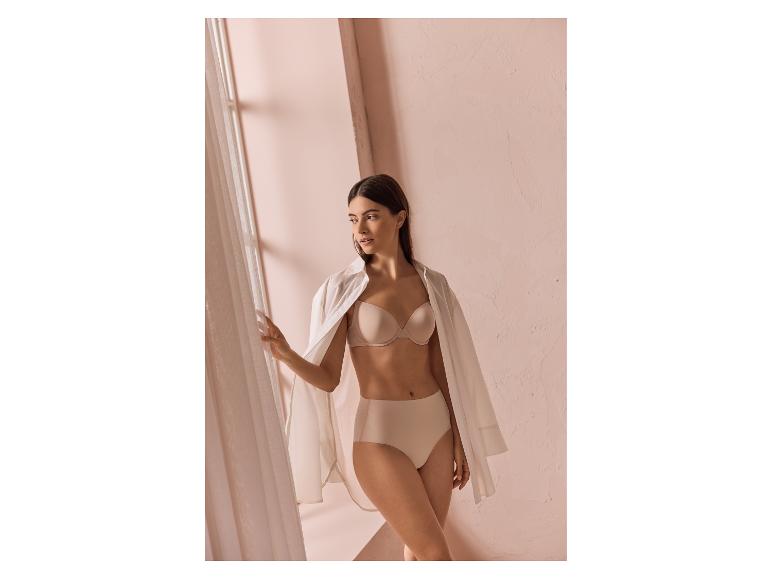 Vrouw in beige beha en slip, met een wit overhemd, staand bij een raam.