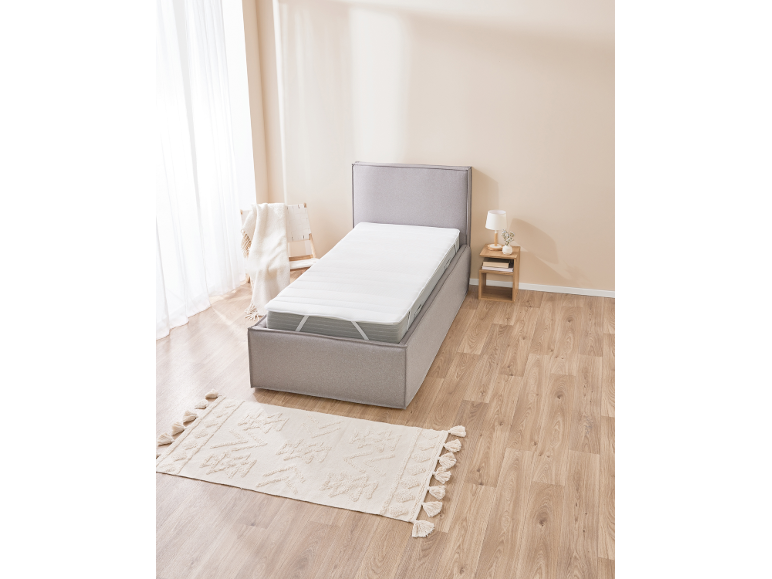 Grijs boxspringbed met wit matras in slaapkamer met houten vloer en beige vloerkleed.