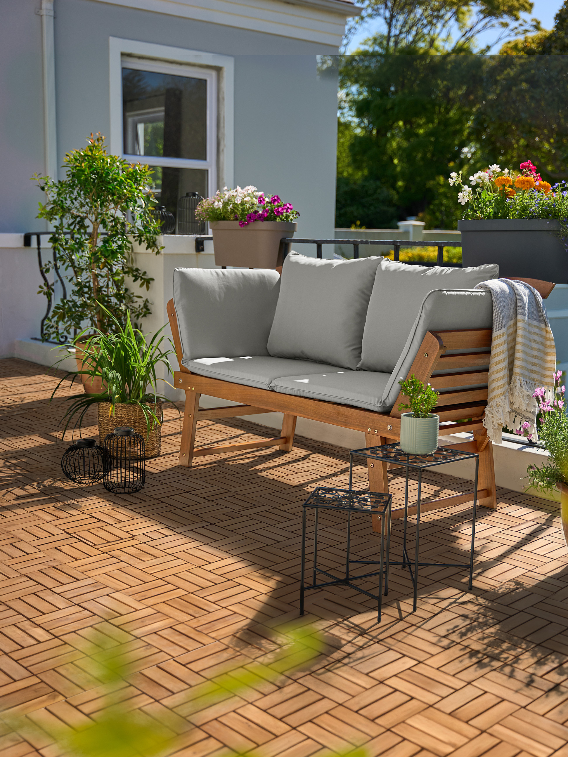 Houten tuinbank met grijze kussens op een balkon met houten vloertegels en planten