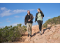 Twee personen in wandelkleding lopen op een rotsachtig pad onder een blauwe lucht.