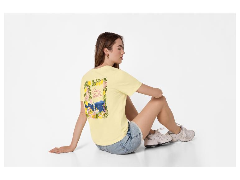 Vrouw in geel T-shirt met 'Ciao Bella'-print en denim shorts, zittend op de grond.