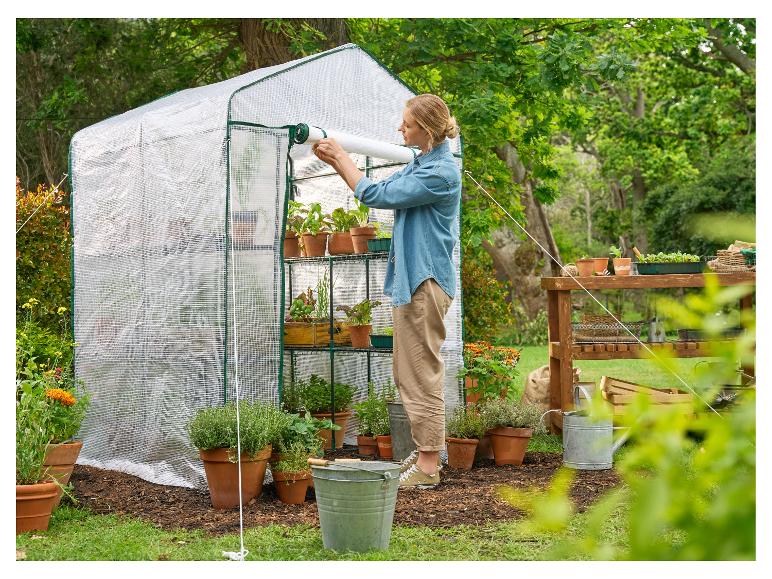 Femme ouvrant une petite serre avec des plantes en pot et des outils de jardinage.