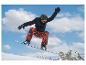 Snowboarder in zwarte jas en oranje broek zweeft in de lucht tegen een blauwe hemel.