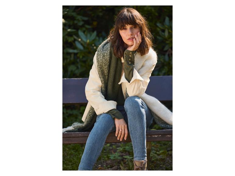 Vrouw in jeans, groene trui en sjaal, zittend op een houten bank.