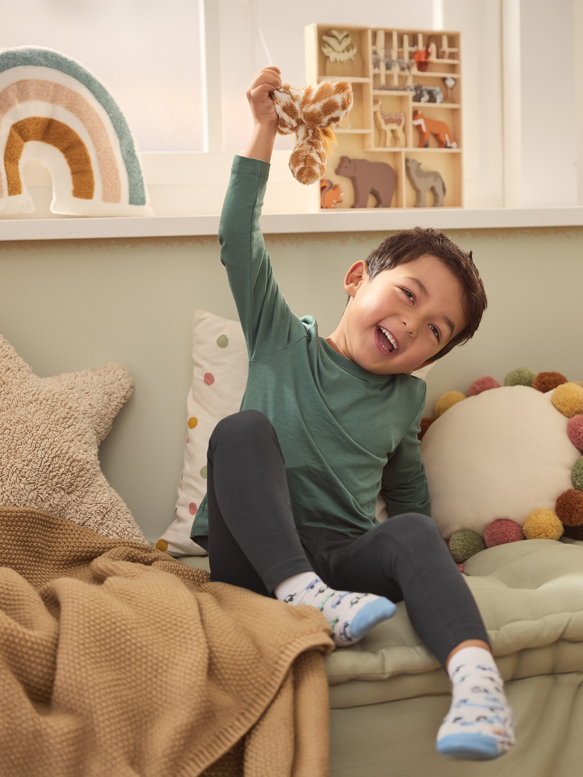 Enfant souriant avec un jouet en peluche girafe, entouré de coussins et d'une couverture.