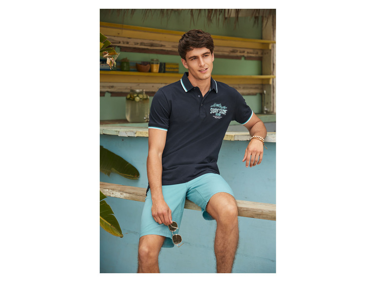 Man in een blauwe polo met 'Surf Shop'-logo en blauwe shorts.