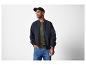 Man in een zwart bomberjack, olijfgroen henley shirt en blauwe jeans.