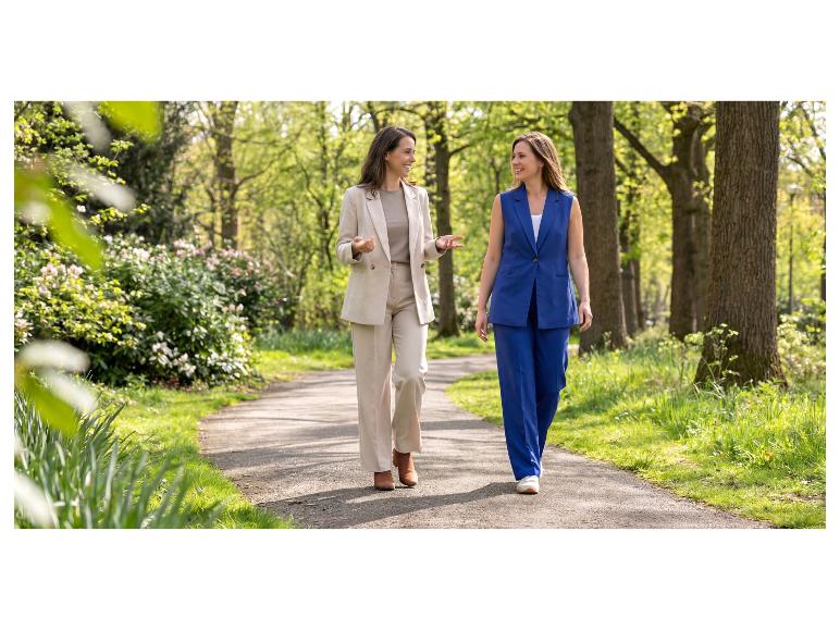 Twee vrouwen in elegante broekpakken lopen en glimlachen in een park.