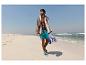Man in turquoise zwemshort en gestreepte handdoek loopt op een zandstrand.