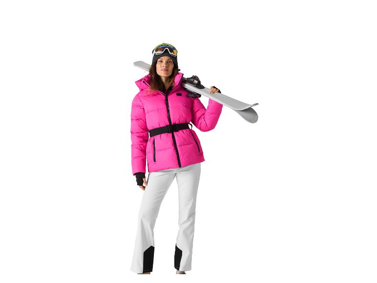 Femme en veste de ski rose, pantalon blanc et lunettes, tenant des skis.