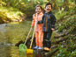Twee kinderen in regenkleding met een schepnet en schep aan de rand van een beek.