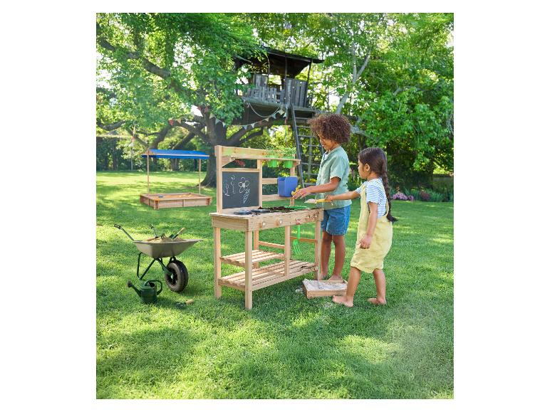Enfants jouant à une table de jardinage en bois avec tableau, avec cabane dans les arbres et bac à sable en arrière-plan.