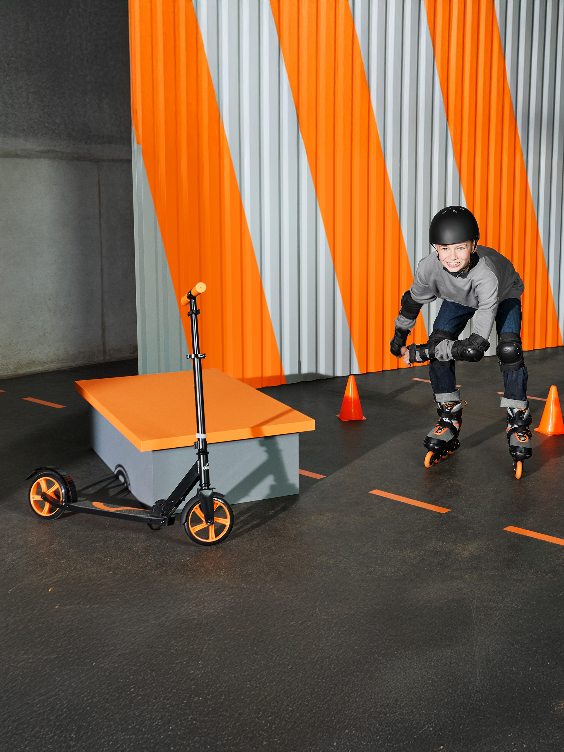 Garçon en rollers et casque, avec une trottinette à côté, dans un espace orange et gris.