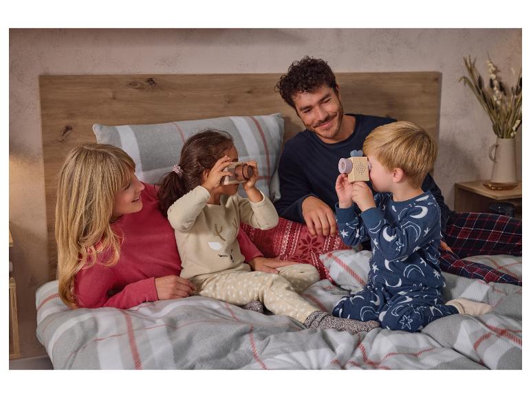 Familie in pyjama op een bed, kinderen spelen met houten speelgoedcamera's.