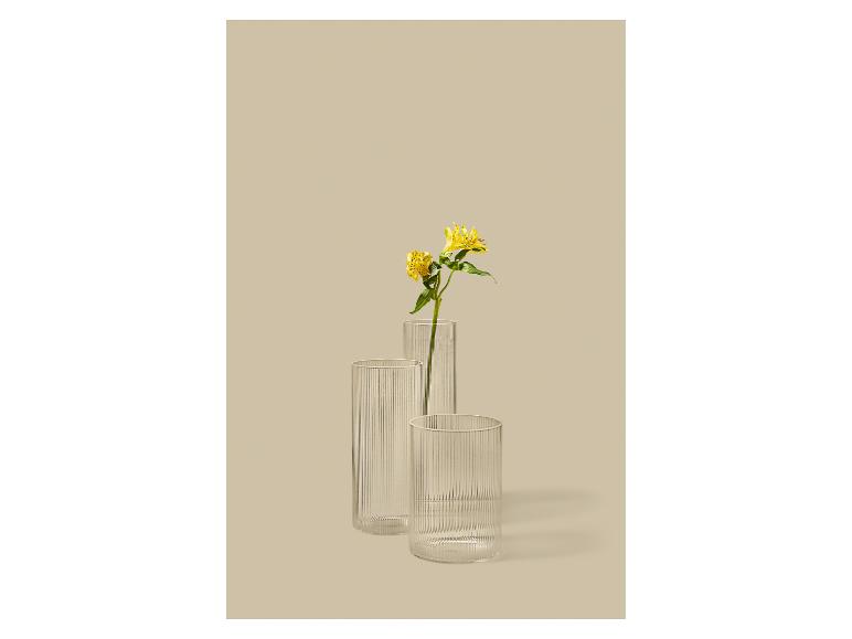 Drie geribbelde glazen vazen, één met gele bloemen, op een beige achtergrond.