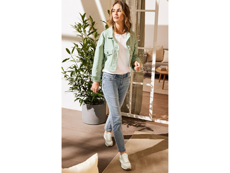 Une femme porte une veste en jean verte, un jean bleu et des baskets blanches.