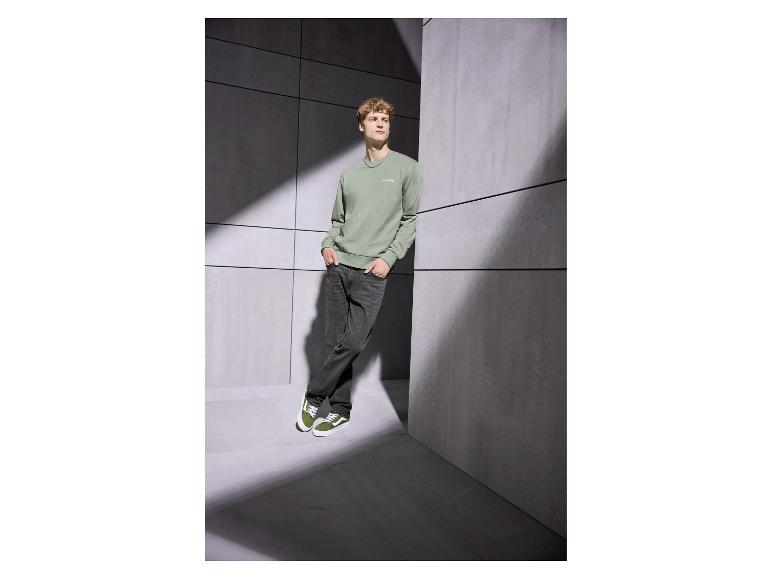 Homme en sweat-shirt vert avec 'Club 1990' et jean foncé, appuyé contre un mur.