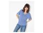 Femme en t-shirt bleu à manches courtes et jean, appuyée contre un mur blanc.