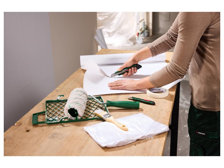 Vrouw snijdt behang met een stanleymes, met schildergereedschap op tafel.