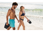 Stel wandelt op het strand, man in zwemshort met slippers, vrouw in bikini met slippers.