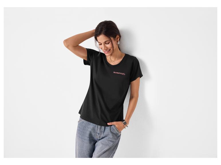 Vrouw in zwart T-shirt met 'BONJOUR'-opdruk en blauwe jeans, lachend, leunend tegen een muur.