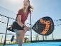 Vrouw speelt padel met een zwart-oranje padelracket op een blauwe baan.