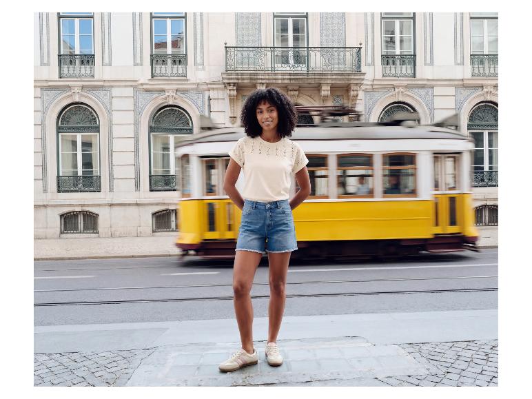 Femme en t-shirt brodé et short en jean devant un tramway jaune en mouvement.