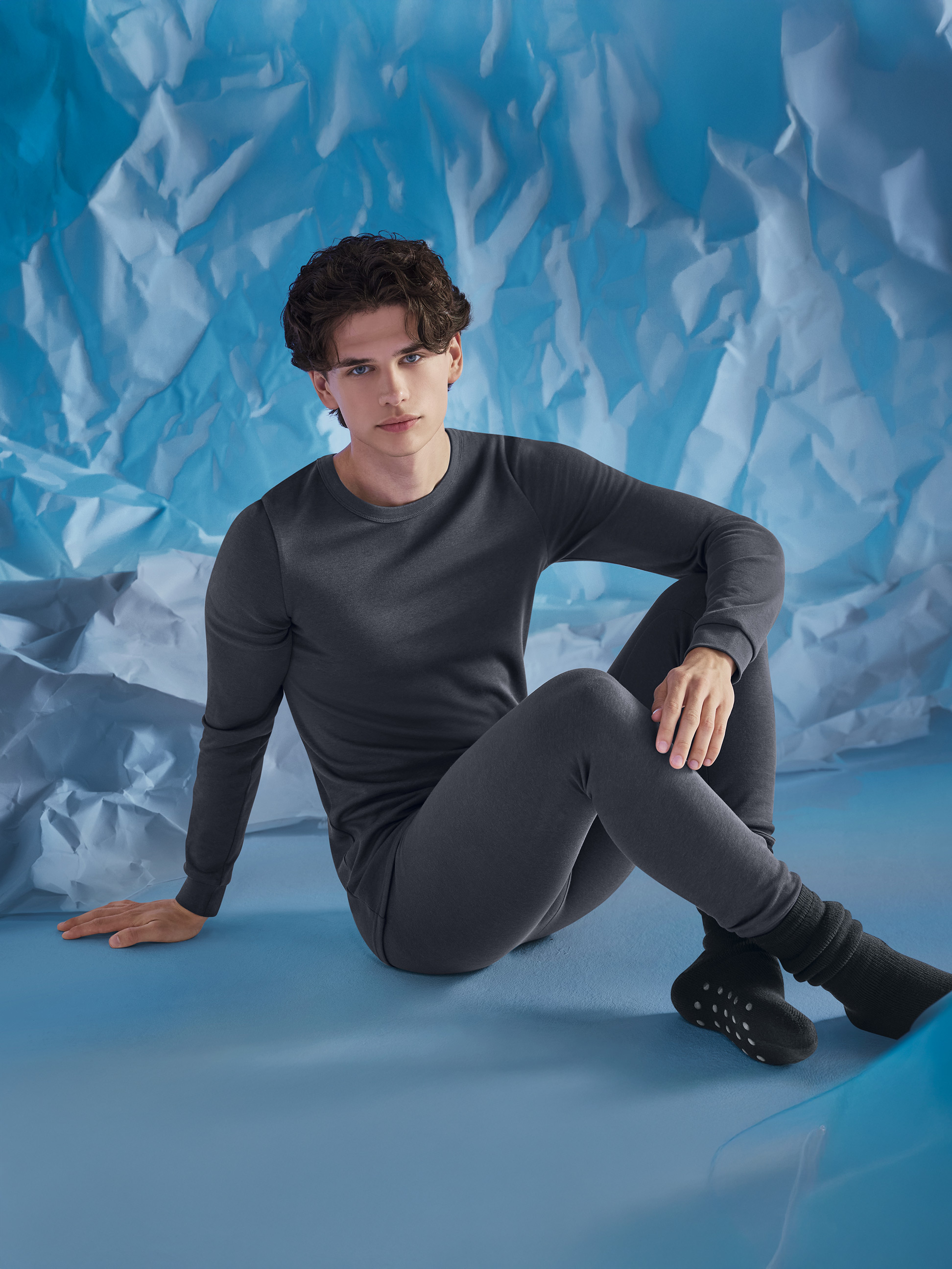 Homme en sous-vêtements thermiques gris, assis sur un fond bleu froissé.