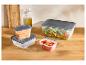 Divers récipients de conservation alimentaire avec couvercles gris, remplis de salade, carottes et pastèque sur une table en bois.