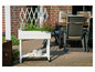 Een witte plantenbak met bloemen en planten, naast tuinstoelen en een tafel.