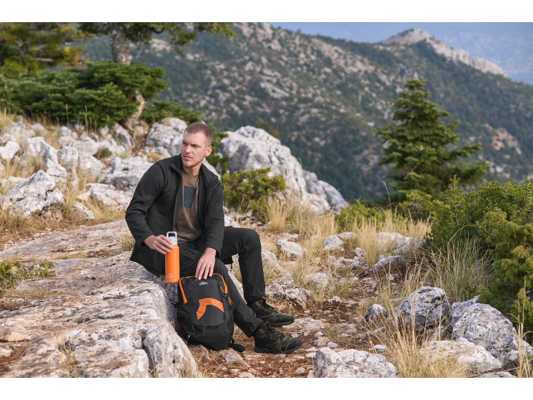Homme en veste polaire et pantalon de trekking assis sur un rocher avec sac à dos et bouteille.