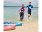 Twee personen in wetsuits rennen het water in, naast een opblaasbaar paddleboard.