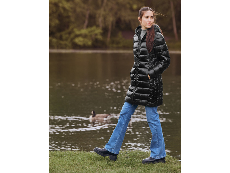 Une femme porte une doudoune noire et un jean bleu, marchant le long d'un lac.
