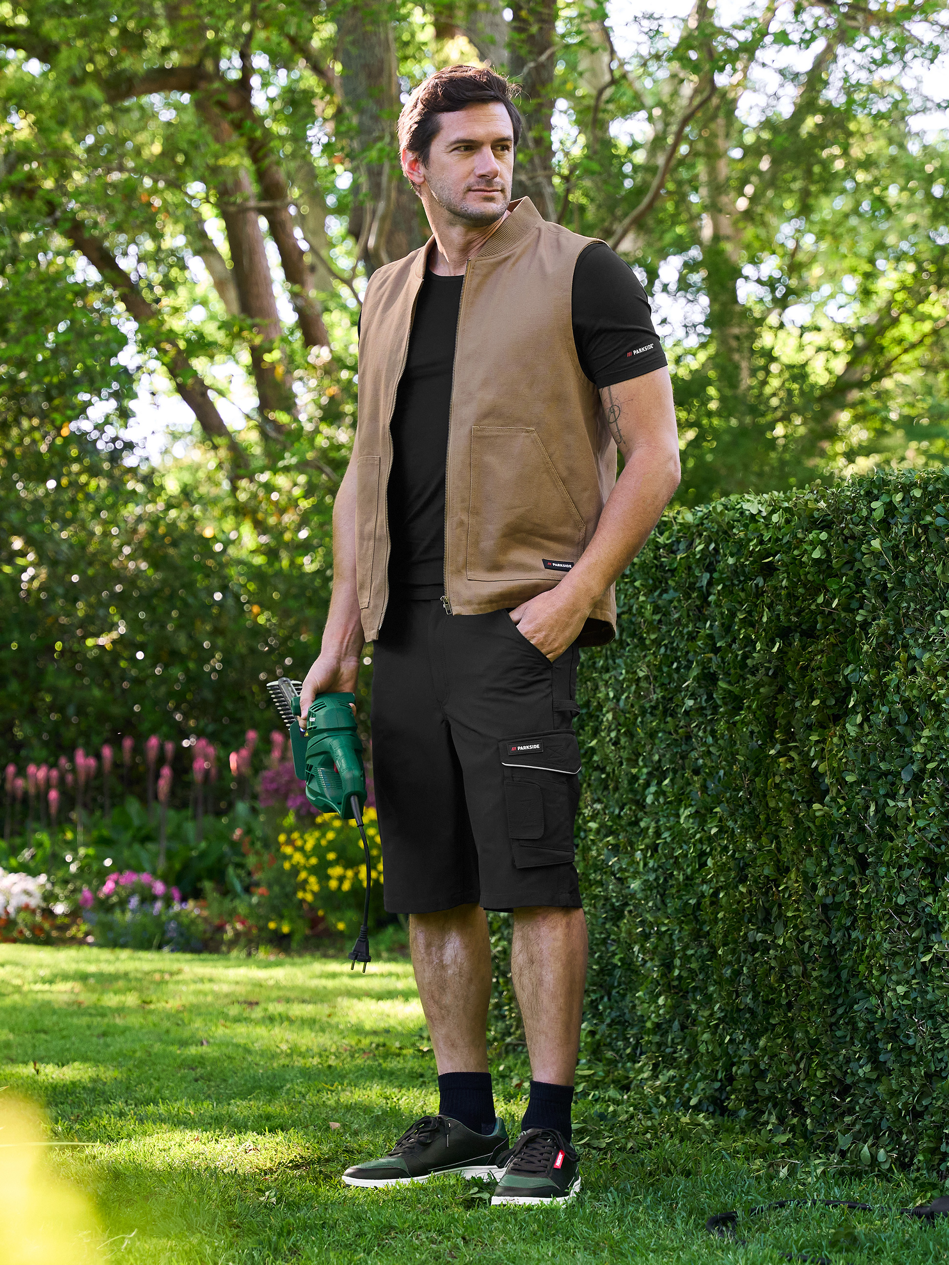 Man in Parkside vest en korte broek, met een elektrische heggenschaar in de tuin.