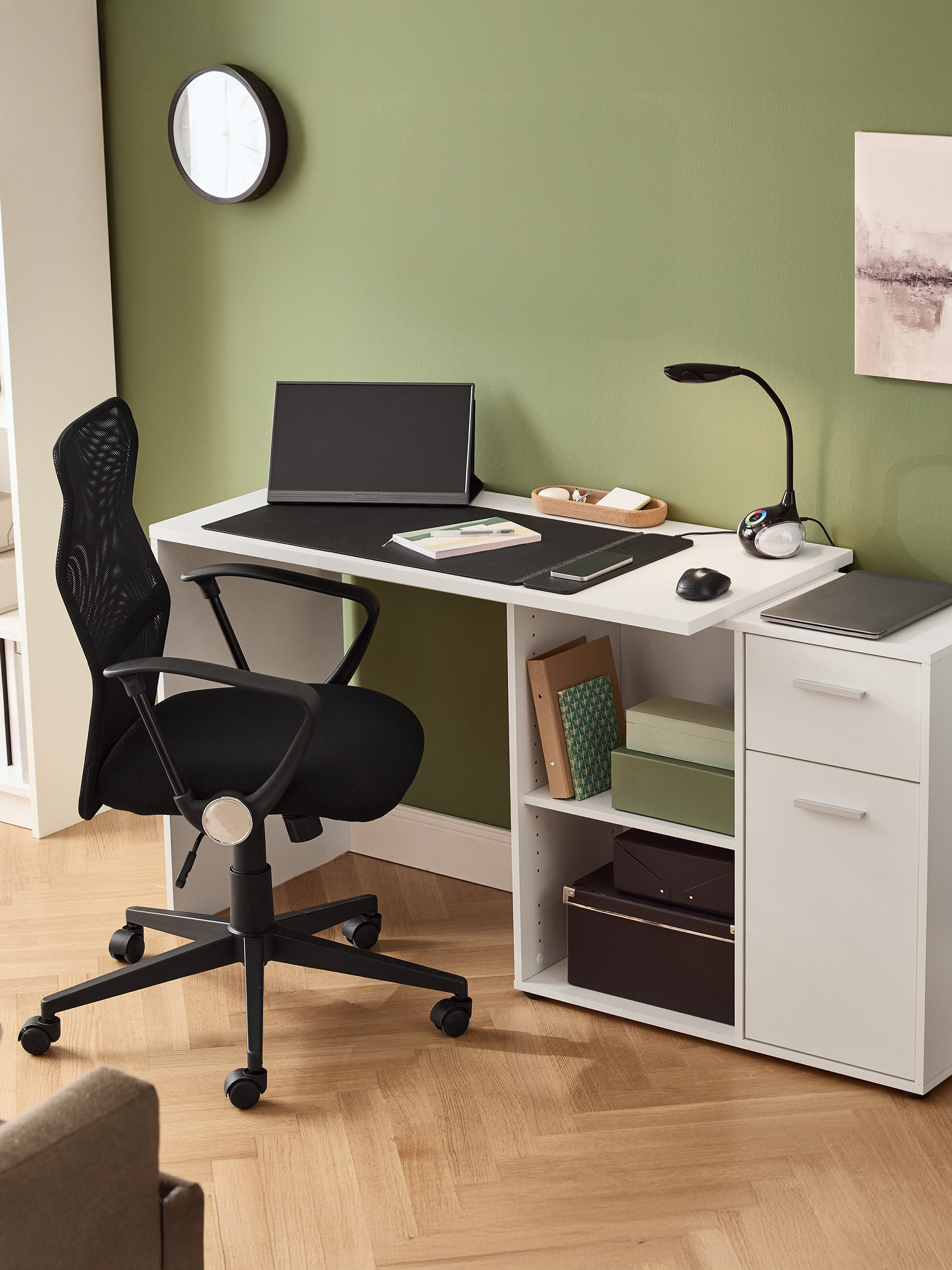 Bureau blanc avec chaise de bureau noire, moniteur, lampe et tiroirs dans une pièce verte.