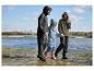 Drie mensen in regenkleding en laarzen lopen langs het water.
