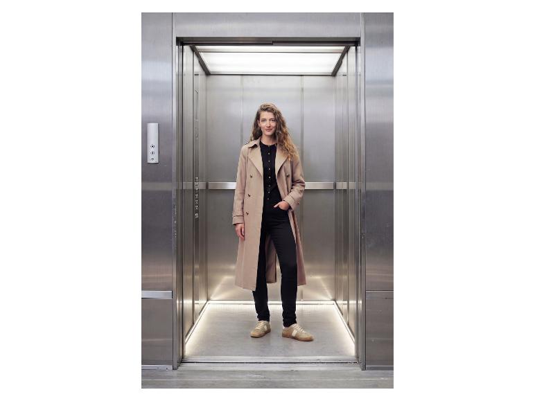 Vrouw in een trenchcoat en zwarte outfit staat in een lift.
